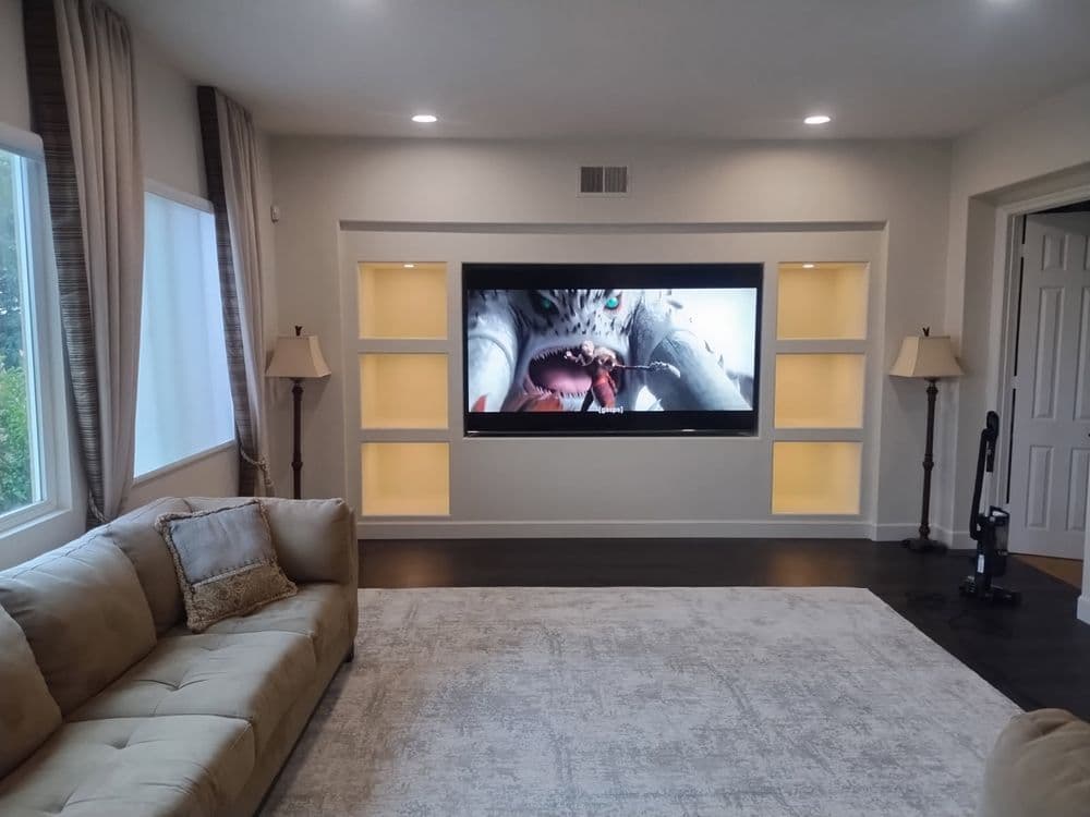 Modern living room with a large TV, beige sofa, and decorative lighting in built-in shelves.